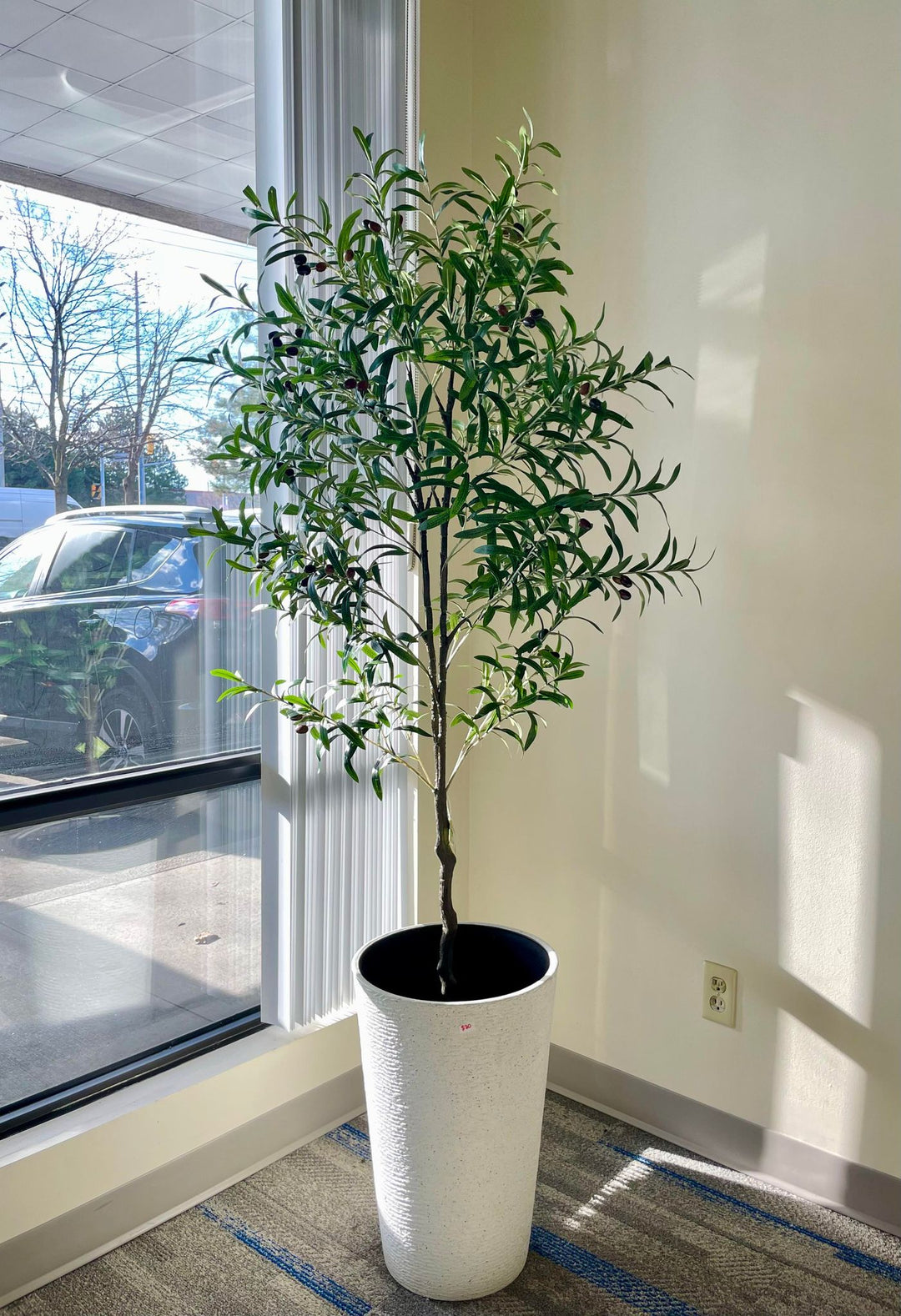 Artificial olive tree with slender green leaves and small black olives in a textured white pot placed near a glass-paneled door in a bright indoor space with wooden flooring