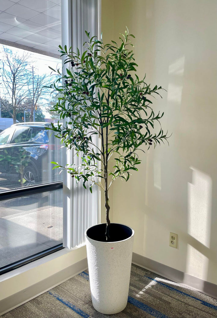 Artificial olive tree with slender green leaves and small black olives in a textured white pot placed near a glass-paneled door in a bright indoor space with wooden flooring