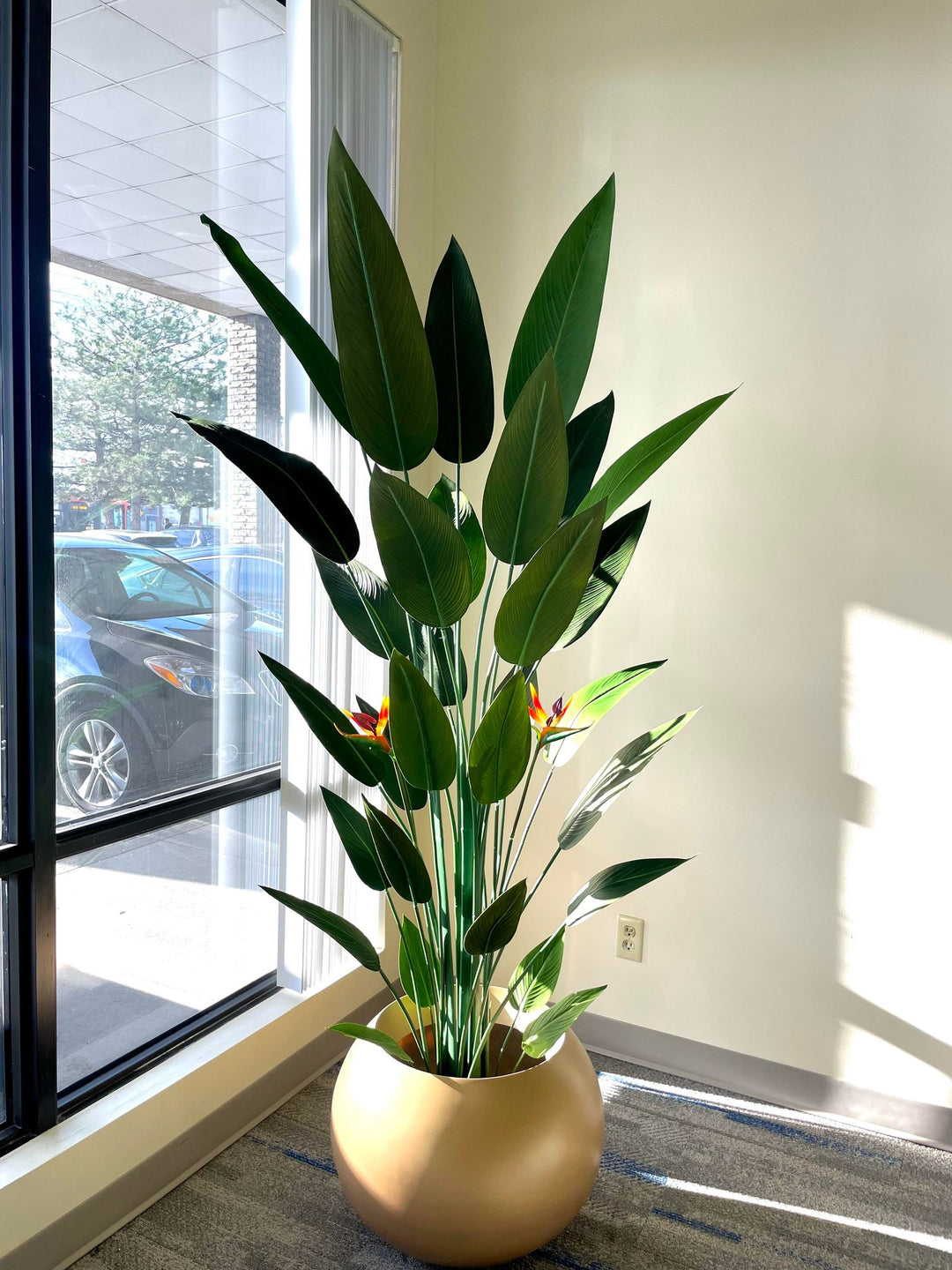 Tall artificial bird of paradise plant with large green leaves and vibrant orange flowers, placed in a round golden pot by a window, with natural light illuminating the plant and casting soft shadows on the floor.