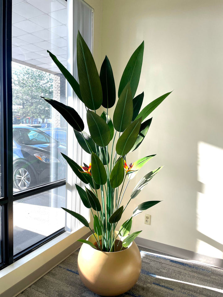 Tall artificial bird of paradise plant with large green leaves and vibrant orange flowers, placed in a round golden pot by a window, with natural light illuminating the plant and casting soft shadows on the floor.
