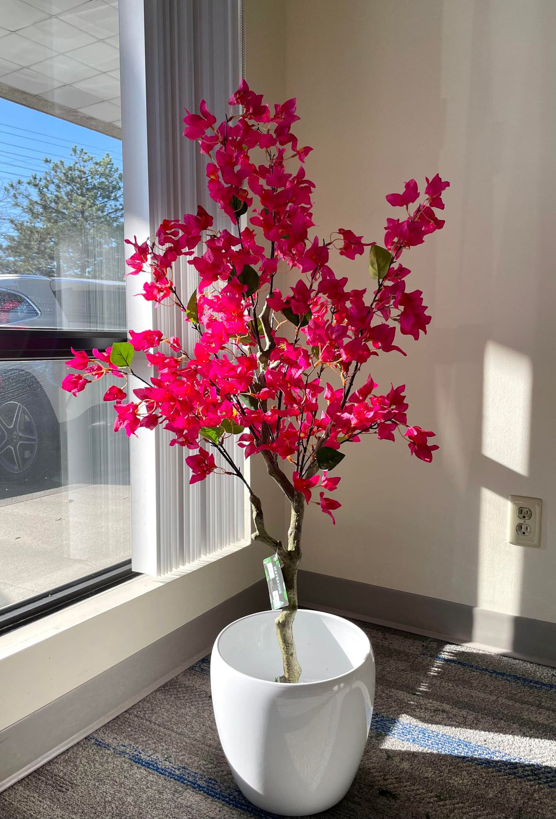 Vibrant artificial pink flowering tree in a white pot, positioned near a large window with natural light streaming in, creating a bright and inviting space