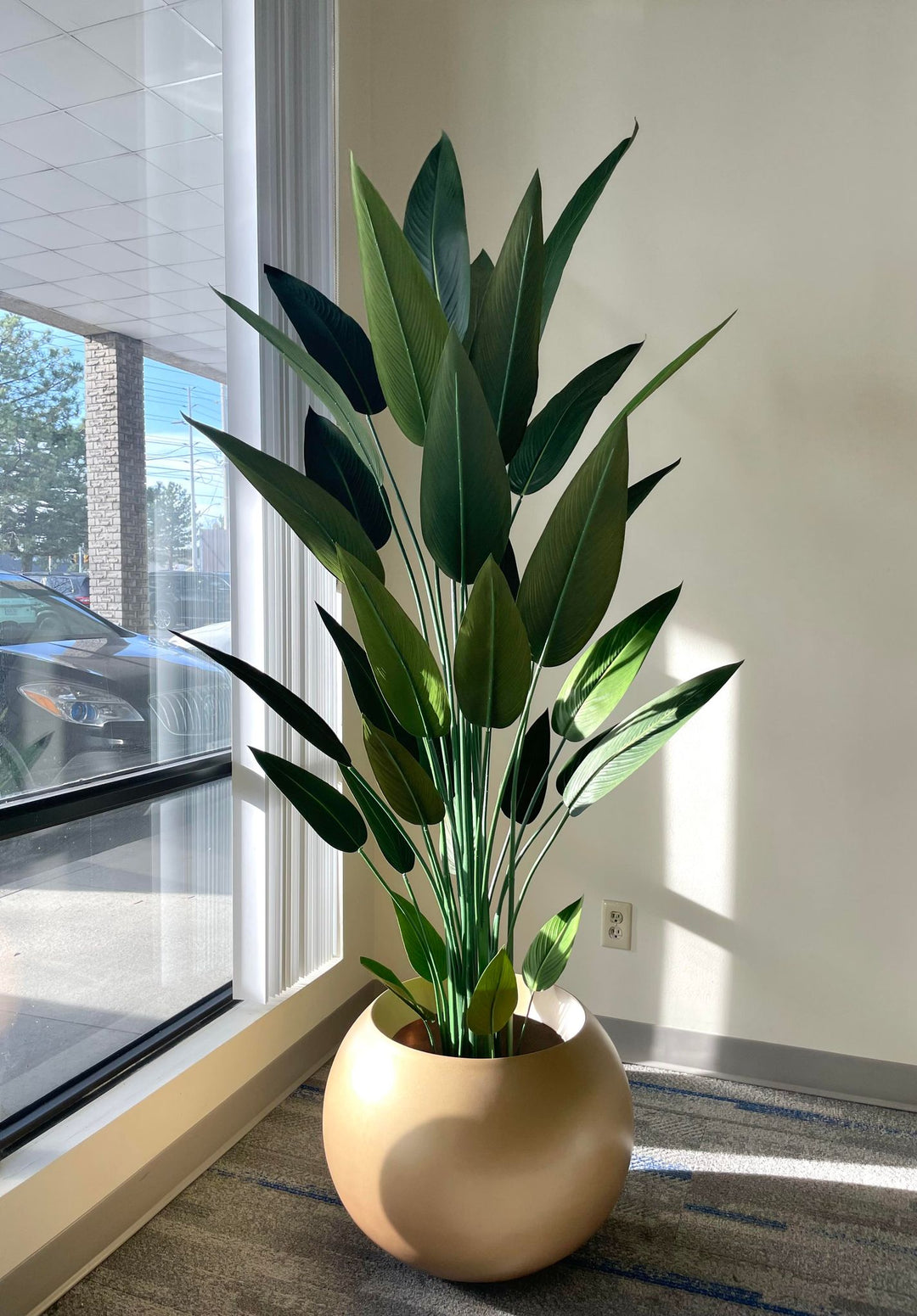 Tall artificial bird of paradise plant with lush green leaves placed in a round, golden planter by a large window, with natural light streaming in, enhancing the vibrant and realistic appearance of the plant.