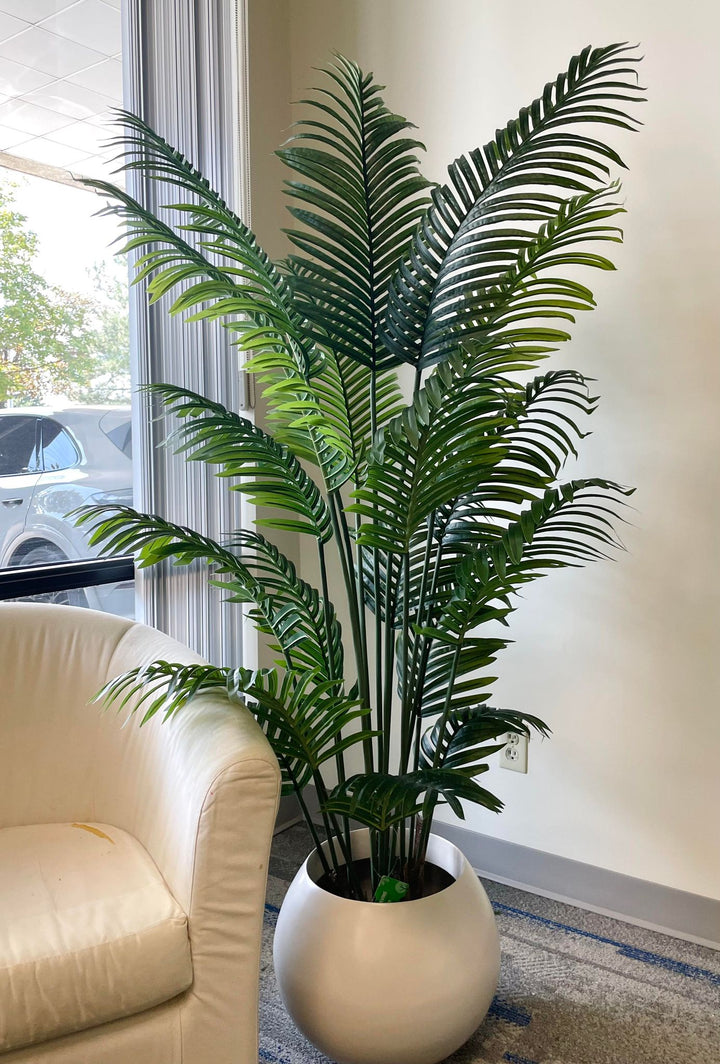 Artificial palm plant in a modern white ceramic pot, placed beside a luxurious white sofa creating a chic and contemporary living room decor.