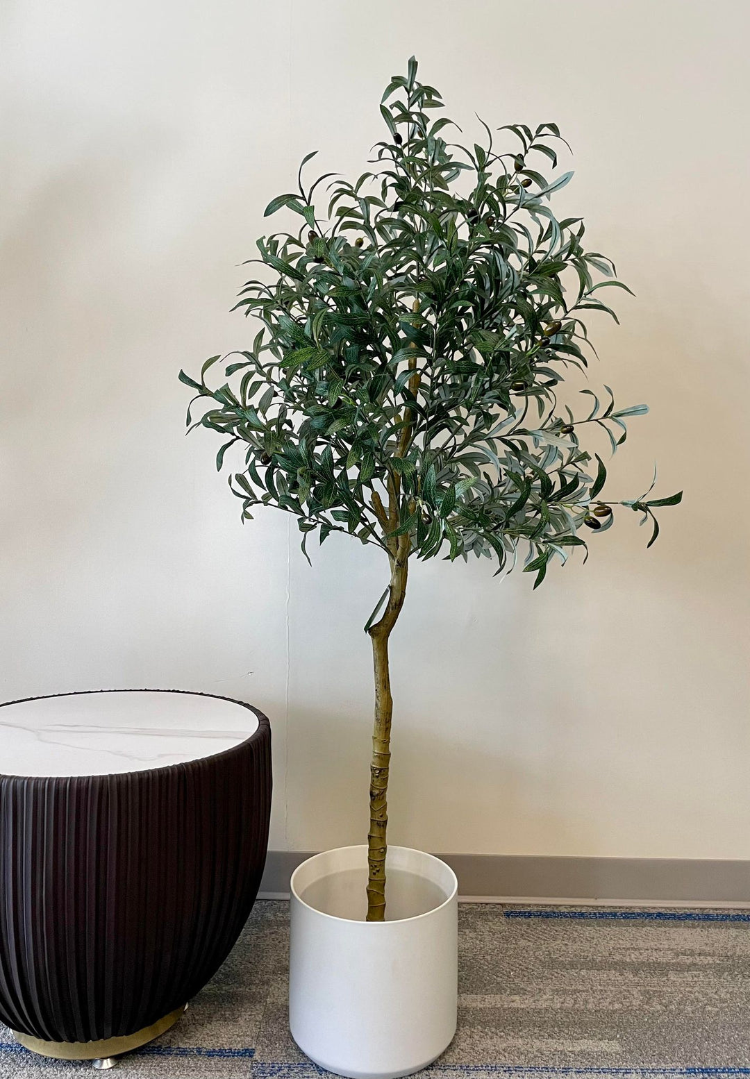 Artificial olive tree with realistic green leaves and black olives in a sleek white pot, positioned next to a modern, dark-colored round side table with a white top, placed on a light-colored carpet in an indoor setting,