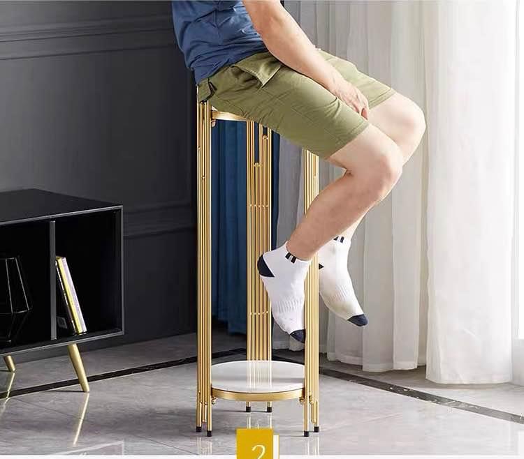 Person sitting on a two-tier gold plant stand with white marble shelves. Frequently asked questions related to this scenario might include: 'Can plant stands hold heavy weights?' 'What materials provide the best stability for furniture?' and 'How much weight can a metal and marble plant stand support?