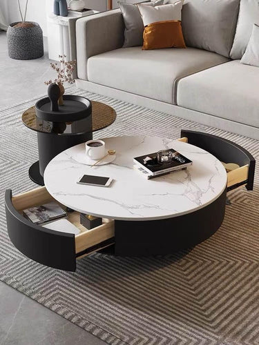 Modern round coffee table with a white marble top and black base featuring built-in storage drawers. Positioned in front of a gray sectional sofa with decorative pillows, the setup includes a black side table and a textured gray area rug, creating a sleek and stylish living room