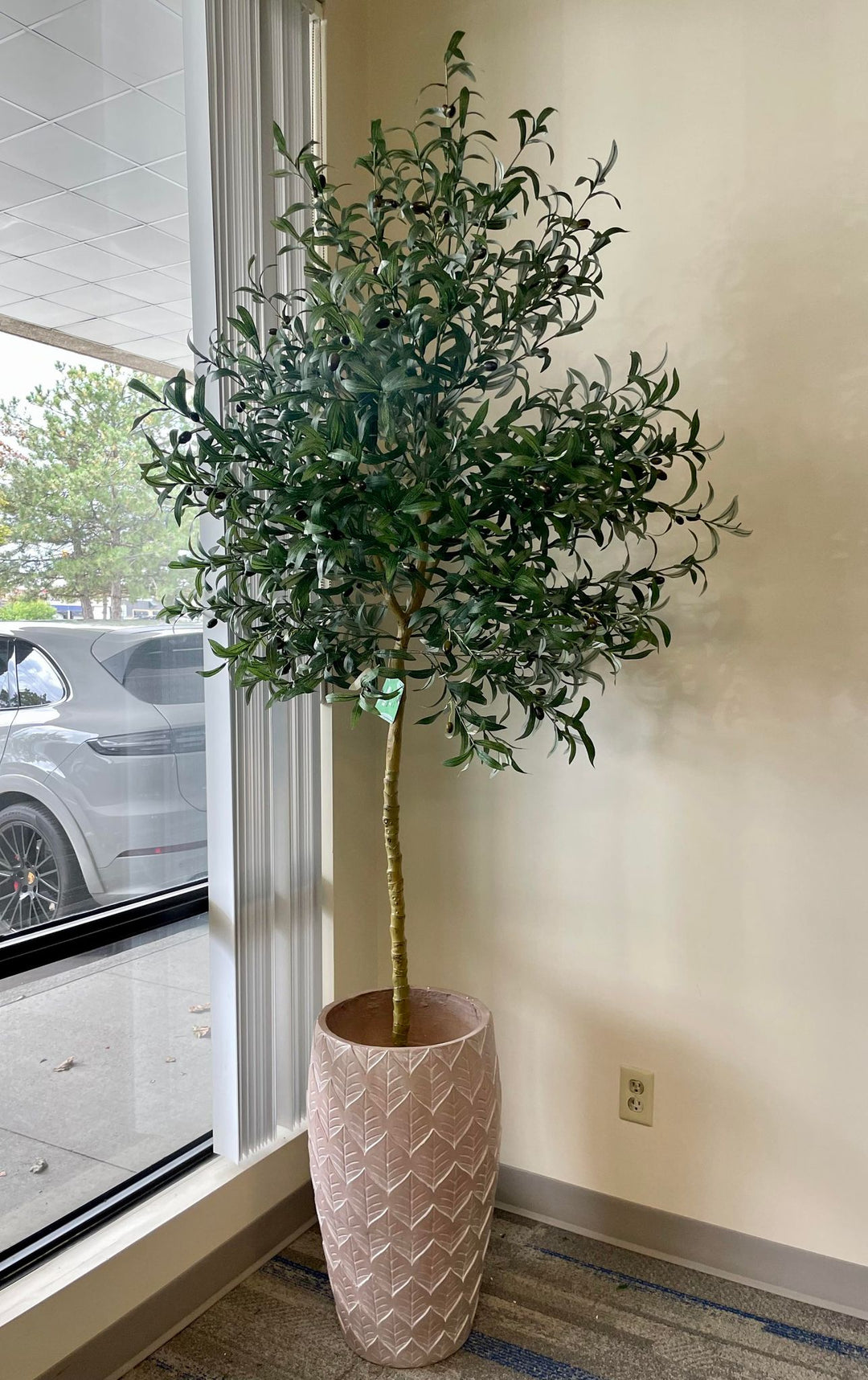 Artificial olive tree placed in a textured, light brown pot with a leaf pattern. The tree is positioned near a window with vertical blinds, filling the corner of a room with a neutral wall color and carpeted floor.