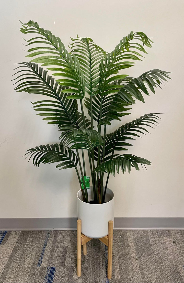 Artificial palm tree in a modern white pot with a wooden stand, displayed against a neutral wall and carpeted floor, ideal for indoor décor.