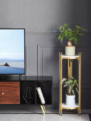 Two-tier gold plant stand with white marble shelves holding potted plants next to a black TV console. Common questions related to this type of furniture include: 'What are the best materials for indoor plant stands?' 'How to style a plant stand in a living room?' and 'Is marble good for furniture durability?