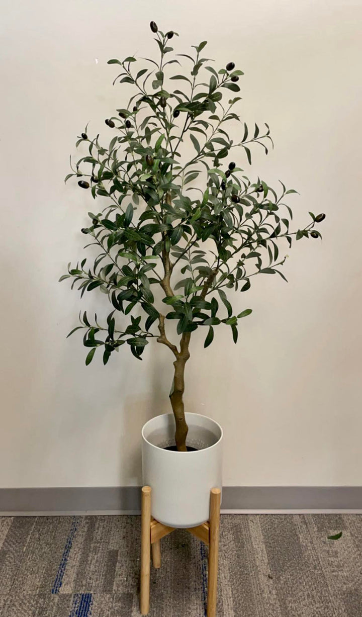 Artificial olive tree in a white planter with a wooden stand, placed against a light-colored wall on a carpeted floor