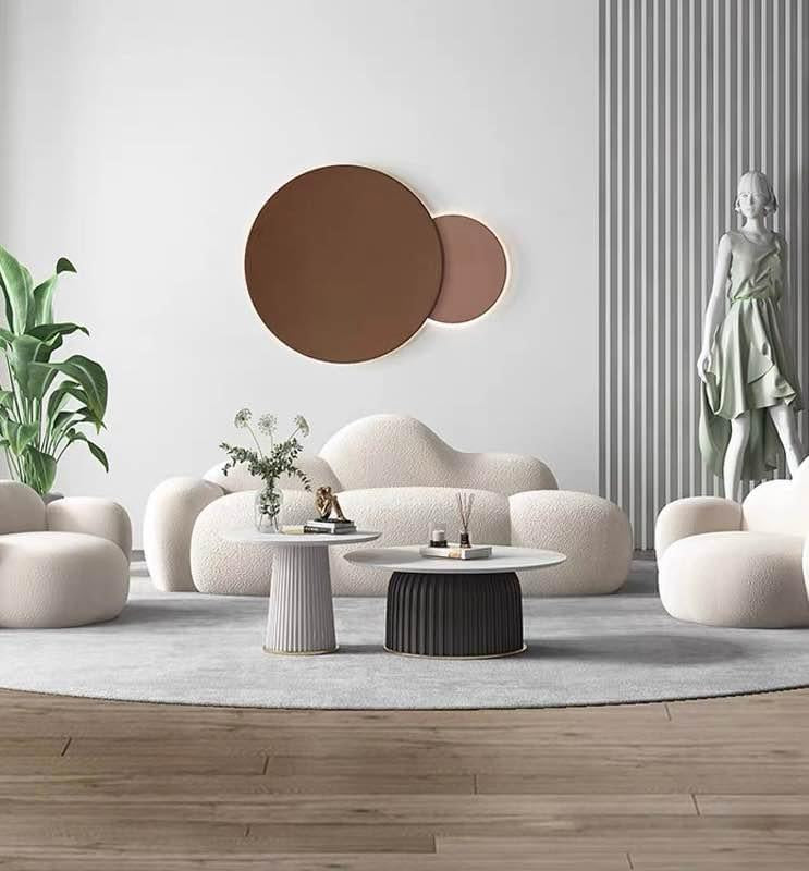 What are the ideal coffee tables for a modern, minimalist living room? This chic space features a cloud-shaped sofa and matching armchairs paired with two round marble coffee tables with ribbed bases. The setup is enhanced by minimalist décor, including a large wall sculpture, indoor plants, and a statue, creating a sophisticated and contemporary design