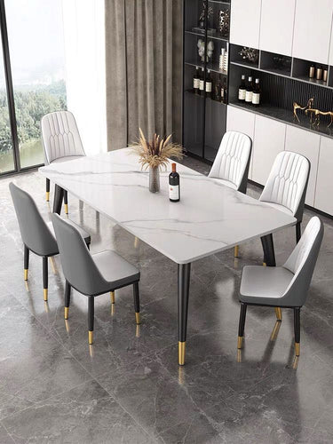 What is the best dining table for a luxury dining room? This elegant dining setup features a large white marble table with sleek black legs and gold accents. The table is surrounded by six upholstered chairs with black and white detailing, creating a sophisticated and modern look. The room is styled with floor-to-ceiling windows and built-in shelving for a luxurious ambiance.