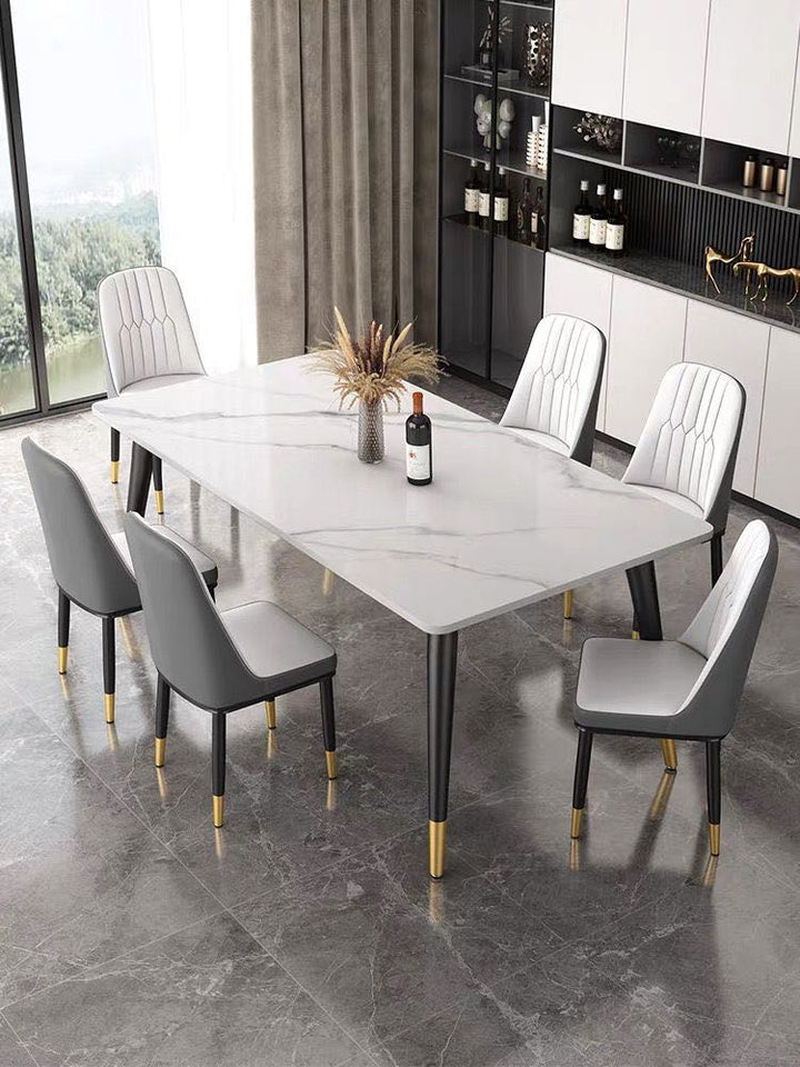 What is the best marble dining table for a luxury home? This modern dining room features a white marble table with black legs and gold accents, surrounded by six upholstered chairs with a gray and white design. The space is complemented by floor-to-ceiling windows and built-in shelving, creating a sophisticated and elegant atmosphere.