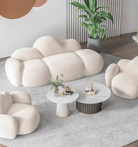 What are the best coffee tables for a cloud couch setup? This modern living room features a plush white cloud-shaped sofa and armchairs paired with two round marble-topped coffee tables. The tables have sleek gold accents and complement the soft, inviting furniture, creating a cozy and stylish atmosphere. A large plant and minimal décor enhance the contemporary design.