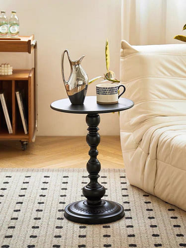 A small, black round side table with a decorative pedestal base is placed next to a light-colored sofa in a cozy living room. On the table are a silver pitcher and a patterned coffee mug. The room has a soft, neutral-toned carpet with a subtle pattern, and natural light streams in from a nearby window, creating a warm and inviting atmosphere.