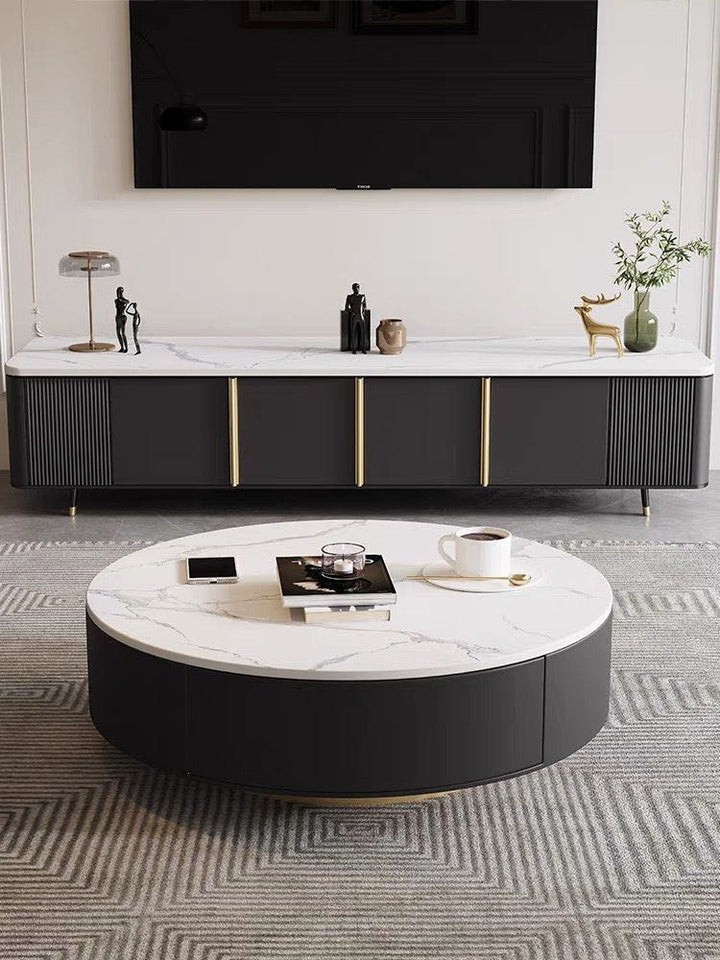 Modern living room with a round coffee table featuring a white marble top and black base. The table is placed on a geometric-patterned rug and in front of a sleek black TV stand with gold accents. Decorative items, including a lamp and sculptures, are arranged on the TV stand, creating a minimalist and sophisticated space