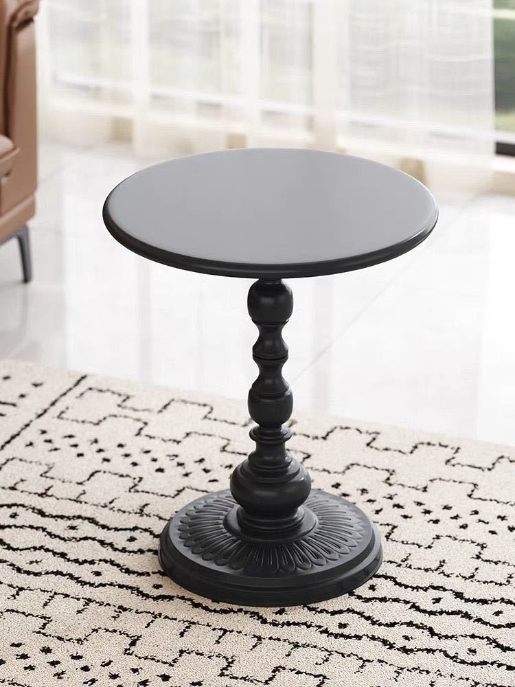 A sleek, round black pedestal side table with an ornate base stand on a white rug with black geometric patterns. The table is placed near a window with sheer curtains, allowing soft natural light to brighten the space, adding a modern and elegant touch to the room.