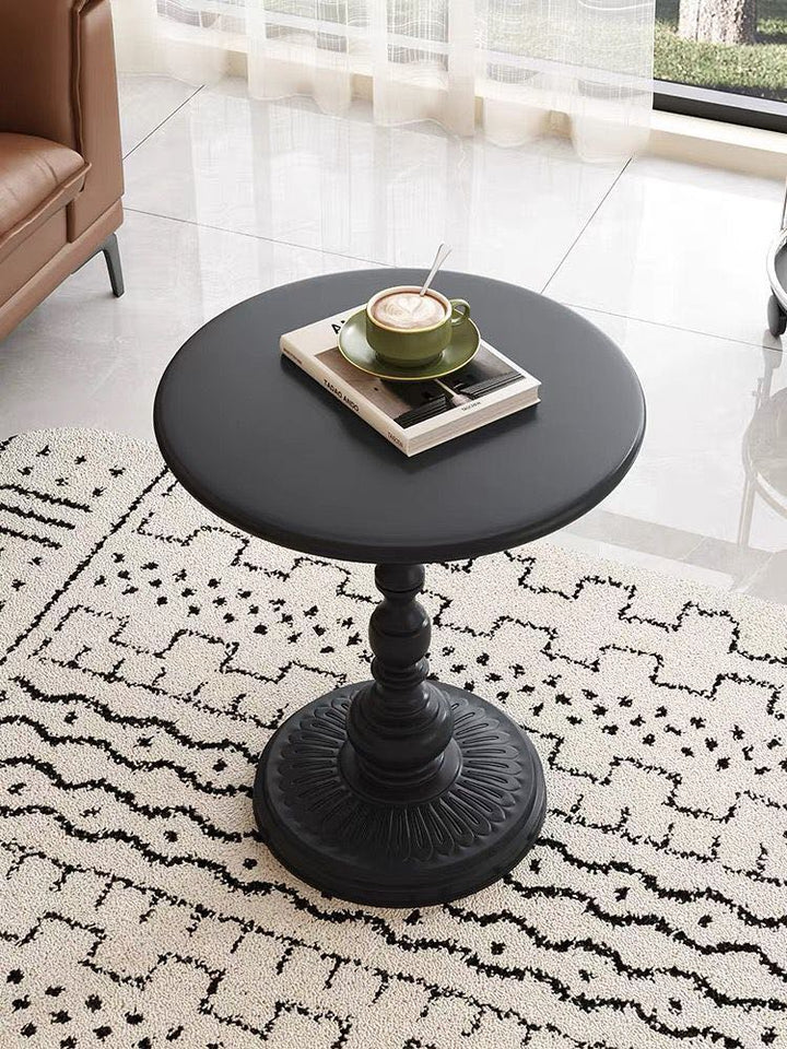 A round, black pedestal coffee table with a sturdy, decorative base sits on a patterned black-and-white rug. A green coffee cup and saucer rest on a book atop the table, creating a cozy, stylish setting near a window with soft, sheer curtains that allow natural light into the room.