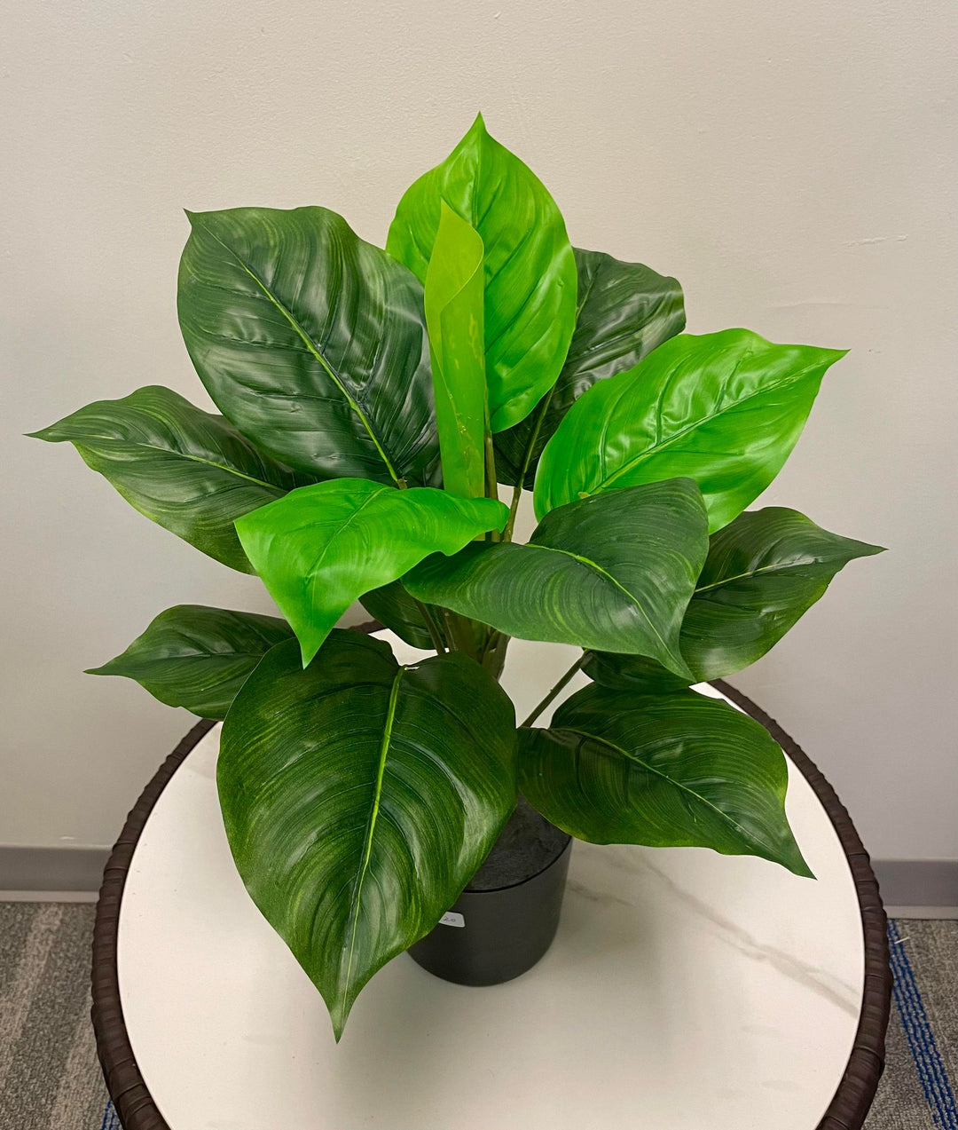 The image shows a potted artificial plant with broad, vibrant green leaves arranged in a compact form. The leaves range in shades from light to dark green, giving the plant a lifelike appearance. It sits atop a round, white tabletop, adding a refreshing pop of greenery to the minimalist space. The plant's natural look makes it an ideal decorative piece for desks or tables in both home and office settings.