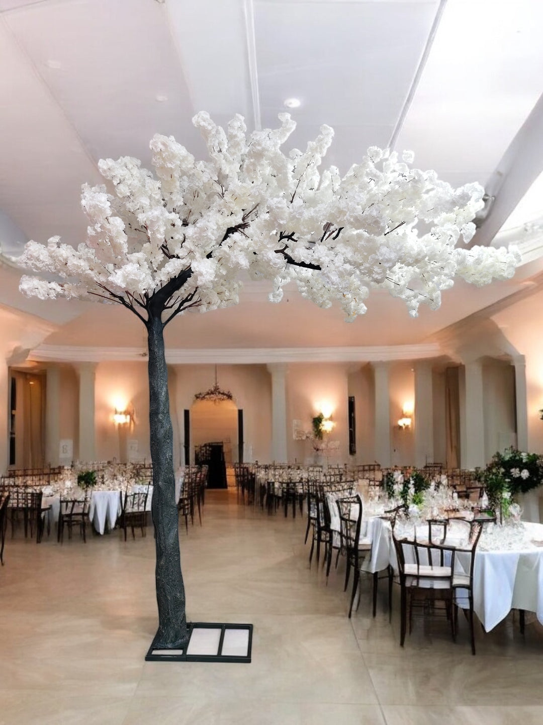 A luxurious indoor wedding venue decorated with a stunning artificial white cherry blossom tree centerpiece. The tree stands tall on a sturdy black base, blending beautifully with the elegant banquet setup, featuring white tablecloths and wooden chairs. How can I secure a decorative tree like this on a polished floor?