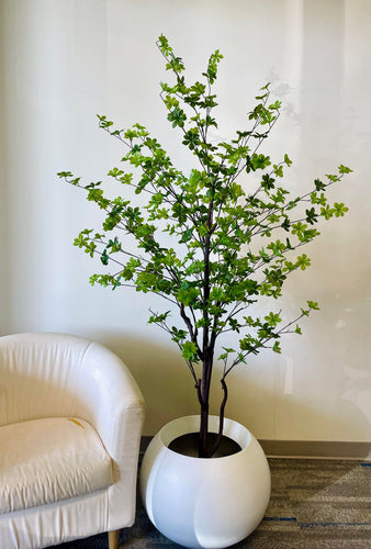 This image features a vibrant artificial plant with lush green leaves, displayed in a sleek, round white planter. The plant is positioned next to a soft, white chair, creating a serene and inviting atmosphere in the space. The bright green leaves are evenly spread across slender branches, adding a refreshing touch of nature to the room. The white planter complements the chair and the neutral background, enhancing the plant's striking green foliage. 