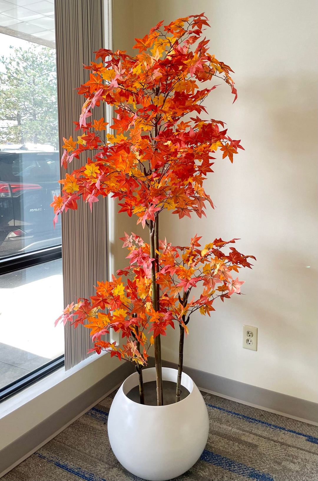 Artificial autumn maple tree with vibrant red, orange, and yellow leaves in a modern white planter, placed near a window in a well-lit indoor setting