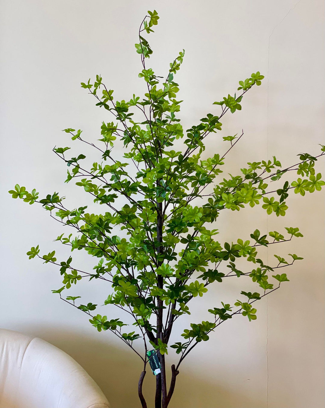 The image shows an artificial plant with lush green leaves arranged in a stylish white pot, placed indoors next to a modern sofa, adding a fresh and natural touch to the room's decor.