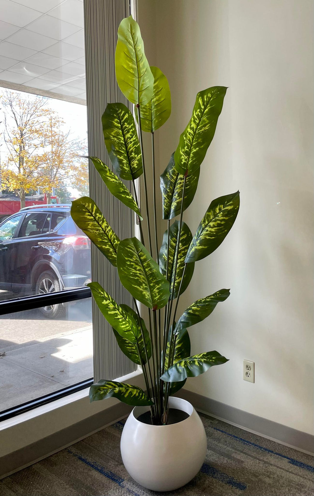 A tall, lush artificial plant with broad green leaves featuring yellow variegation is elegantly placed in a sleek, round white pot. Positioned next to a cozy white armchair, this setup adds a fresh and vibrant touch to the room's decor.