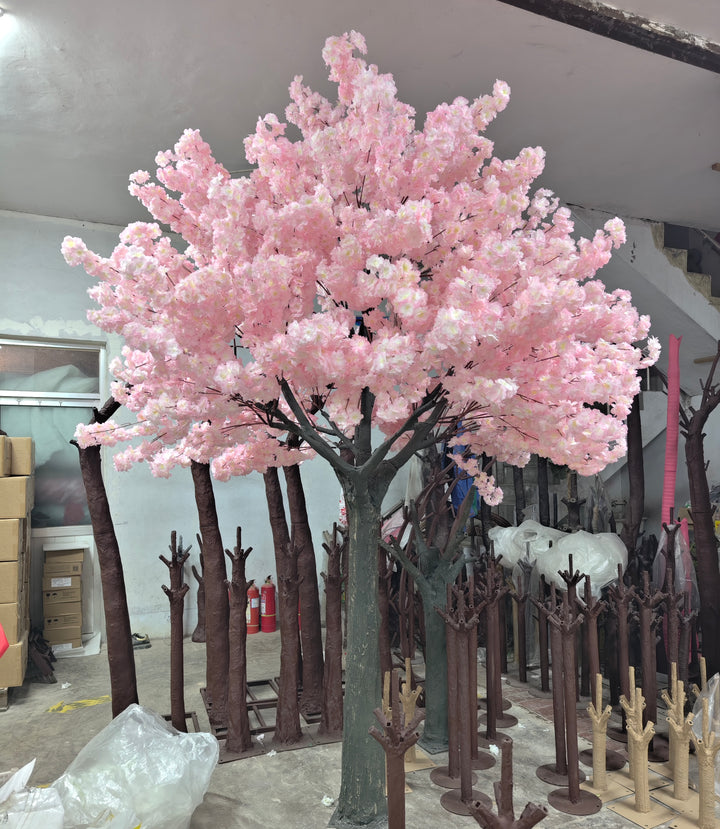 A realistic artificial pink cherry blossom tree surrounded by various tree trunks and bases in a workshop setting.