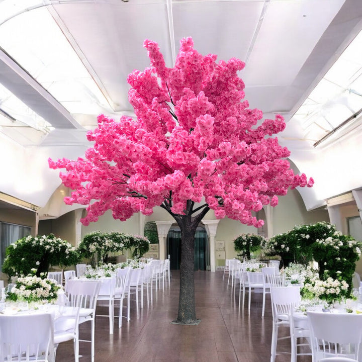 A vibrant artificial pink blossom tree standing in a spacious, elegant hall with white chairs and floral decorations, ideal for weddings or luxury events.