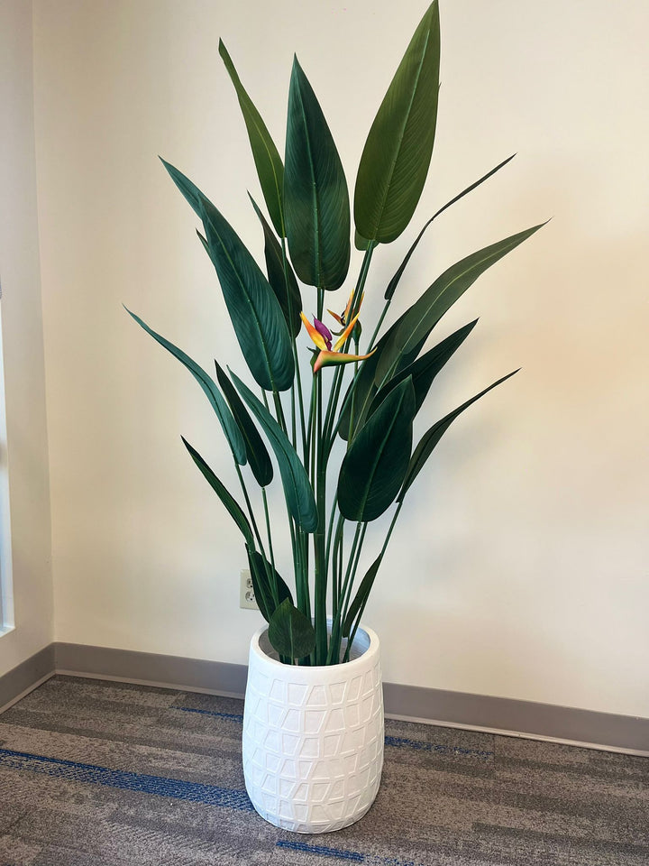 White geometric-textured planter holding a tall artificial bird of paradise plant with vibrant green leaves and orange flowers, placed indoors on a carpeted floor. Is this planter lightweight and easy to reposition?