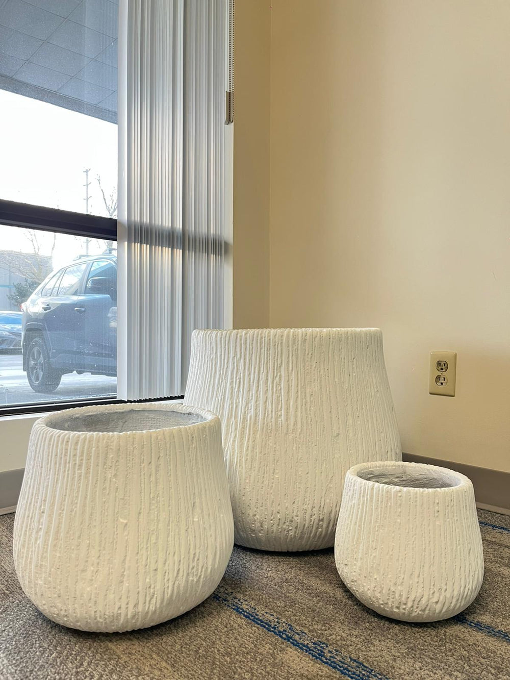 Set of three modern white textured ceramic planters in varying sizes placed indoors near a window with natural light. The planters have a ribbed design, perfect for adding elegance to indoor spaces. Do these planters come with drainage holes?