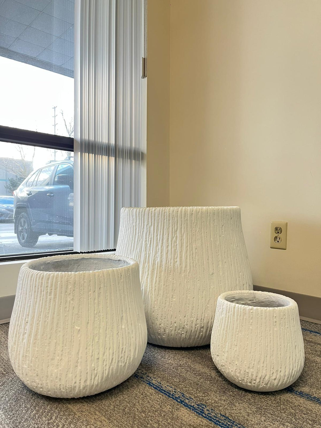 Set of three modern white textured ceramic planters in varying sizes placed indoors near a window with natural light. The planters have a ribbed design, perfect for adding elegance to indoor spaces. Do these planters come with drainage holes?