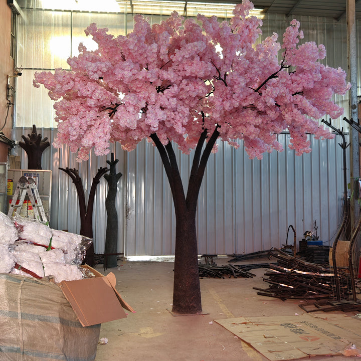Artificial tree with enchanting pink blossoms, displayed in a workshop alongside floral materials and assembly tools, showcasing its preparation for wedding or event rentals. Are these trees easy to assemble and disassemble for event setups?