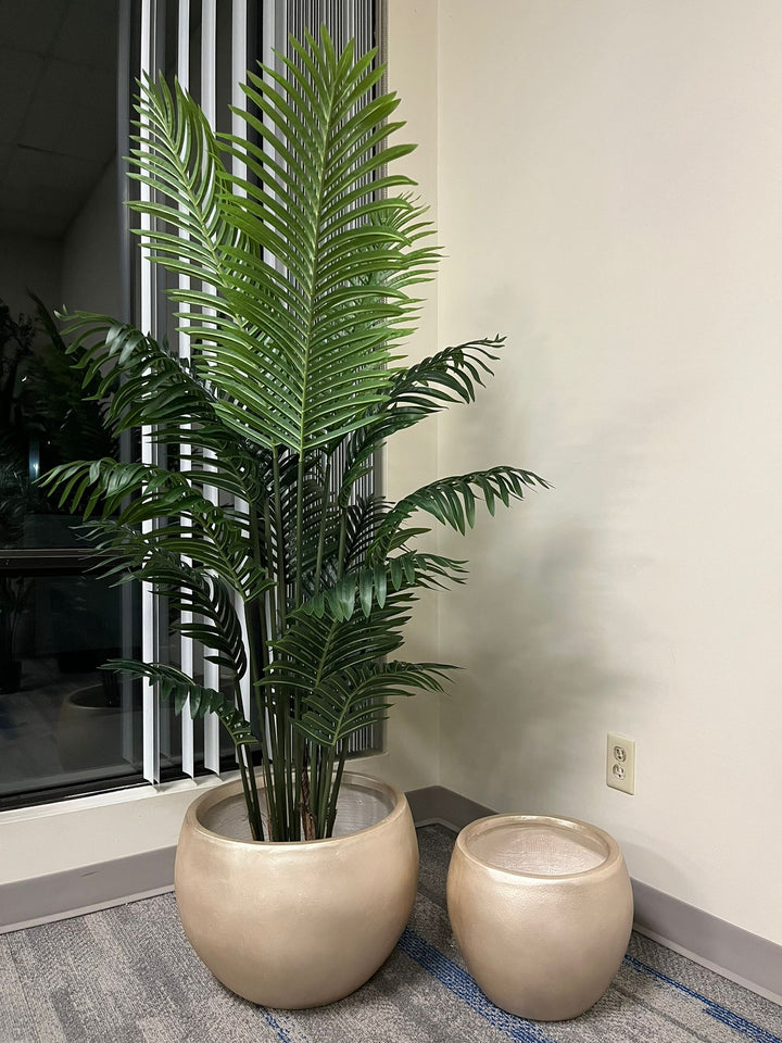 Set of two gold metallic round planters with a smooth finish, one holding a tall artificial palm plant with lush green leaves, placed indoors near a window. Are these planters rust-resistant and suitable for outdoor use?