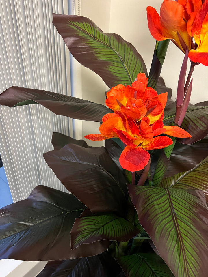 Close-up view of an artificial tropical canna lily plant with vibrant orange flowers and lush, dark green leaves with detailed veining, showcasing the realistic texture and color.