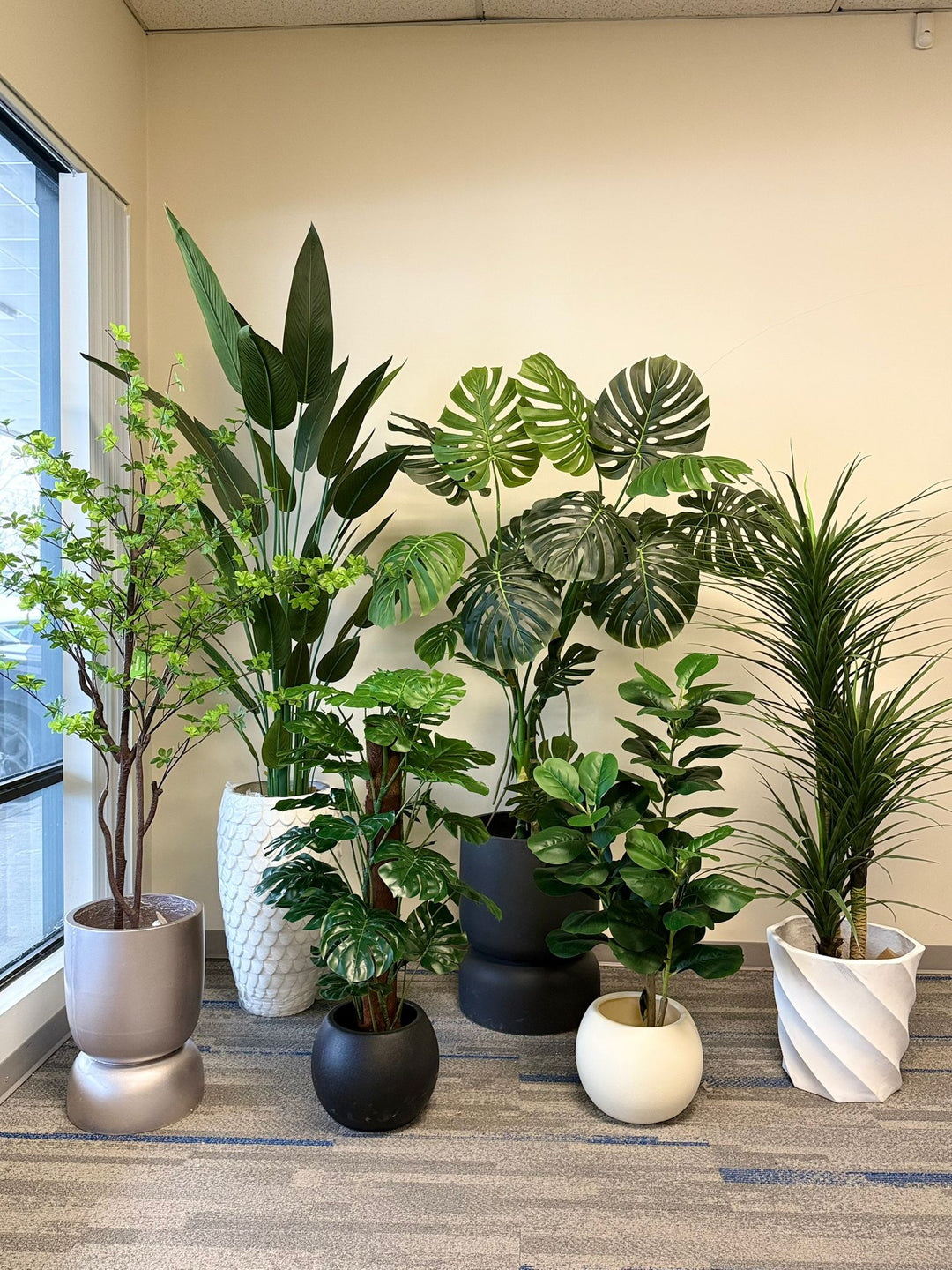 Collection of potted artificial tropical plants on a carpeted floor near a window