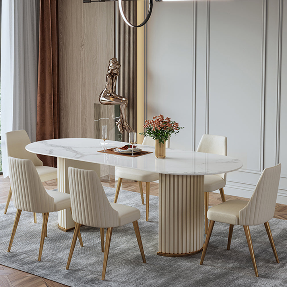 Luxurious round marble-top dining table with golden legs, paired with plush cream-upholstered chairs featuring vertical channel tufting and gold-toned legs, set on a soft gray rug in an elegant, light-filled room.