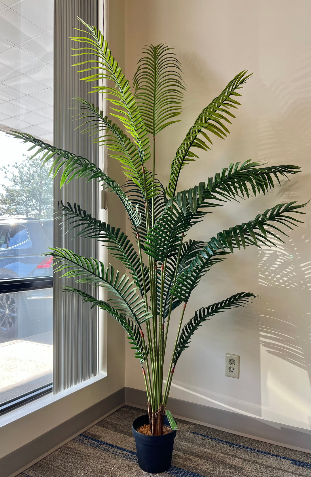 Artificial palm tree in a black pot by a window, with lush green fronds creating natural shadows, perfect for home or office decor.