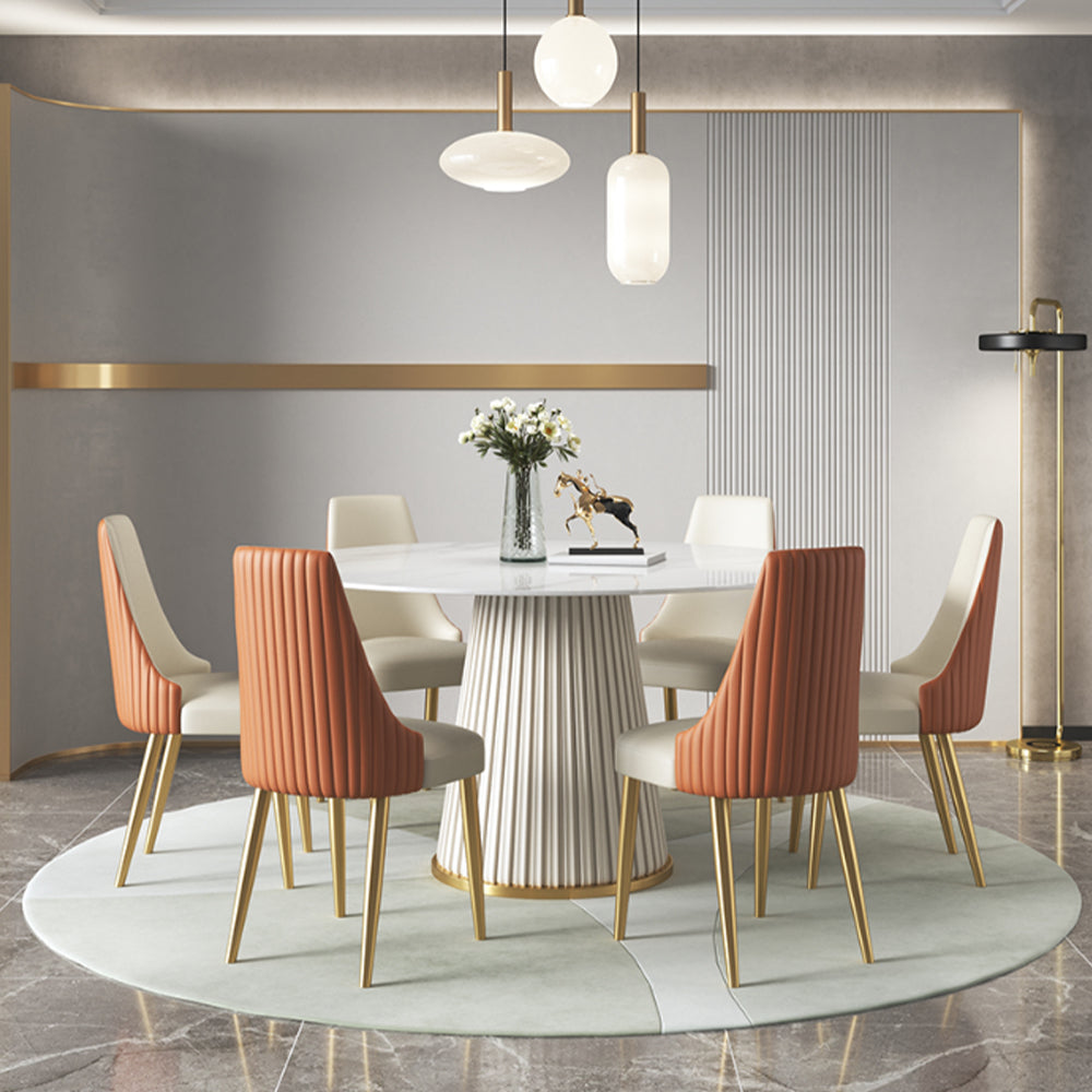 Elegant Round Dining Table with Pleated Base and White Top, Surrounded by Orange and White Upholstered Chairs with Gold Legs in a Modern Dining Room Setting with Pendant Lights and Decorative Accents