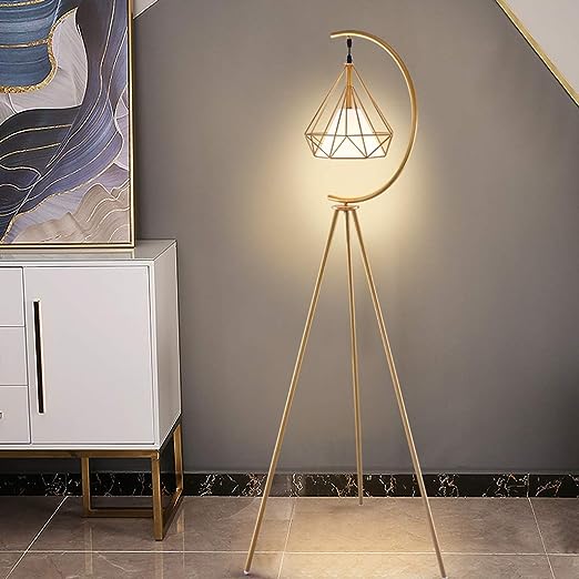 A modern gold floor lamp with a geometric lampshade and crescent-shaped frame, placed next to a white and gold console. The lamp illuminates a soft glow against a dark grey wall, complementing the elegant, minimalist decor of the space.