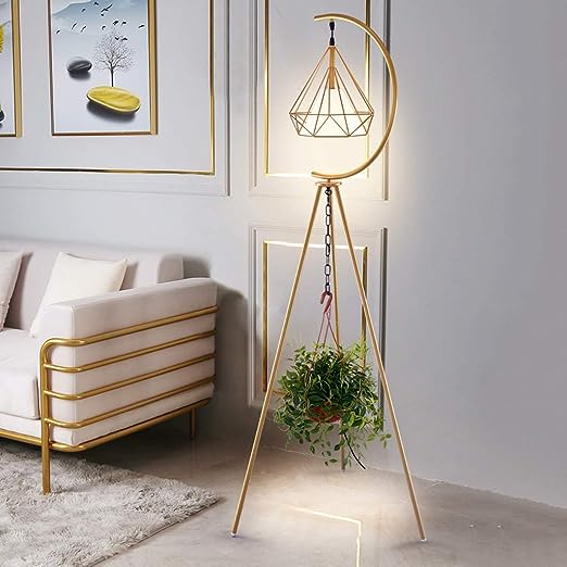 A modern golden floor lamp with a crescent-shaped frame supporting a geometric lampshade, standing beside a minimalist white and gold-accented sofa. A potted plant hangs from the center of the lamp, adding a touch of greenery. The room has light-colored walls adorned with framed artwork and subtle gold detailing.