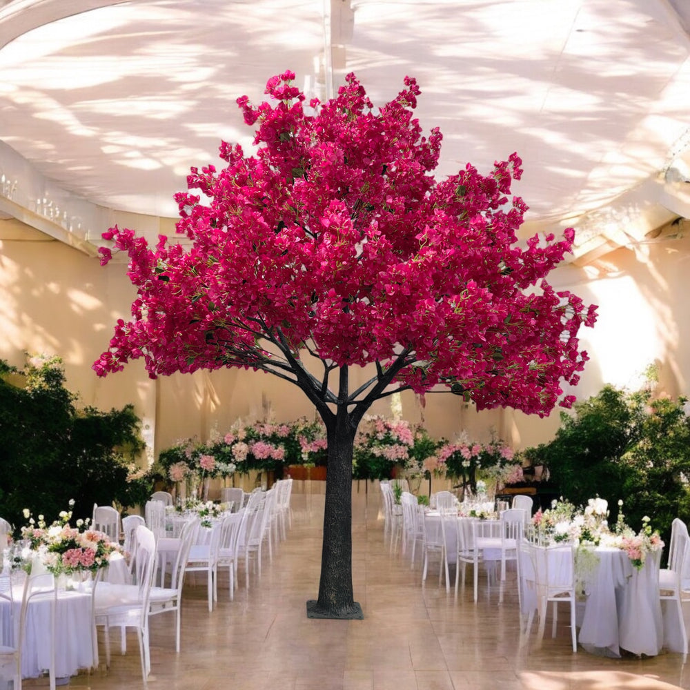 Stunning artificial bougainvillea tree with bright pink flowers, serving as a focal point in an elegant indoor wedding venue. The space is decorated with white chairs and floral-adorned tables, surrounded by lush greenery, and softly lit with natural light filtering through the draped ceiling.