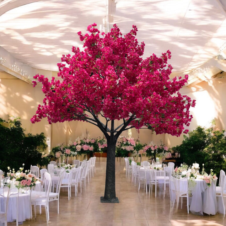 Stunning artificial bougainvillea tree with bright pink flowers, serving as a focal point in an elegant indoor wedding venue. The space is decorated with white chairs and floral-adorned tables, surrounded by lush greenery, and softly lit with natural light filtering through the draped ceiling.