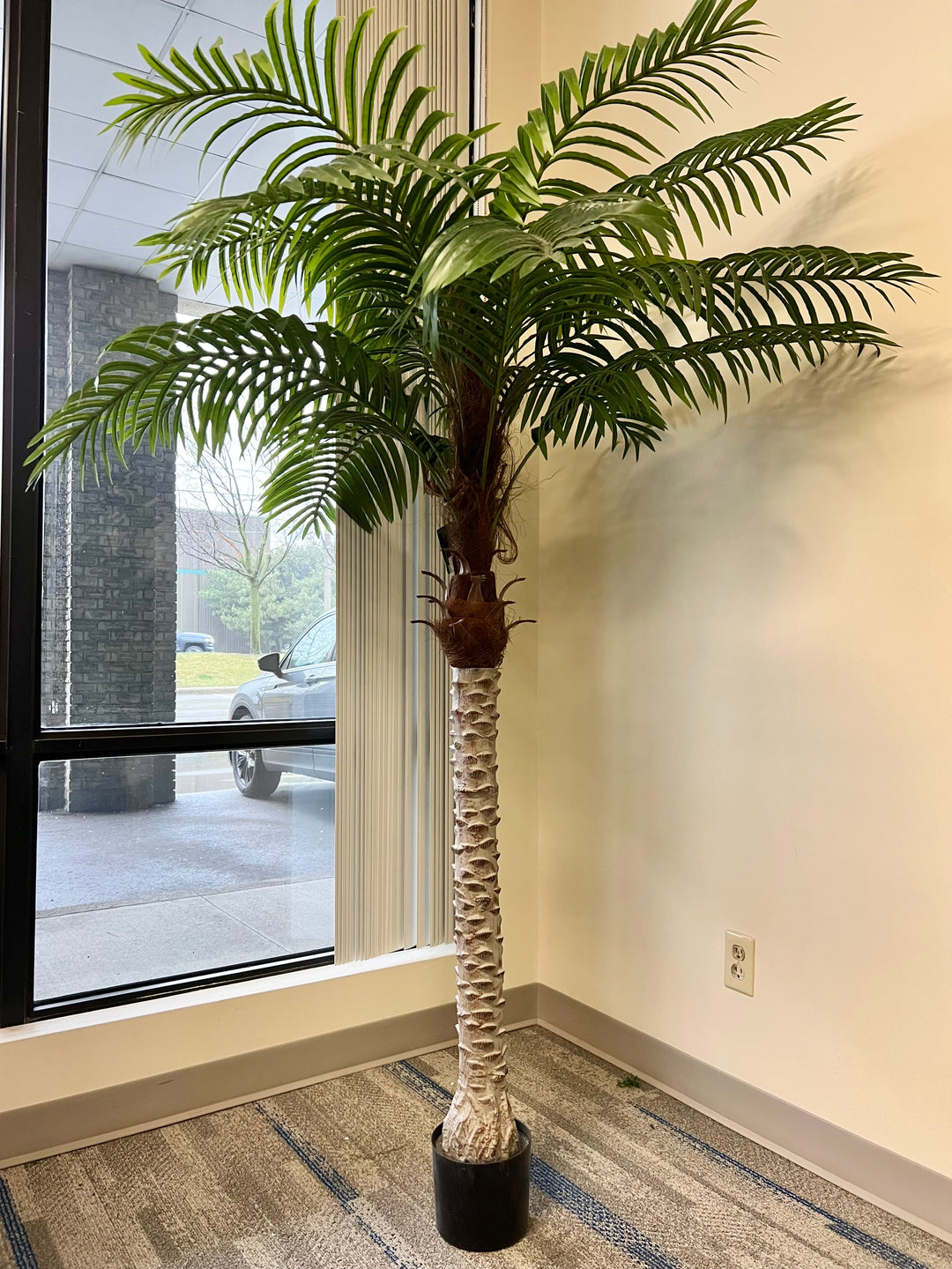 A large artificial palm tree in a stylish office corner by the window, adding a natural touch to a professional setting.
