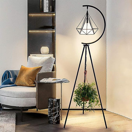 Modern black metal tripod stand with a geometric pendant lamp, featuring a unique crescent moon design, standing in a cozy living room corner. A small potted plant hangs from the center of the stand, adding a touch of greenery to the stylish, minimalist setup.