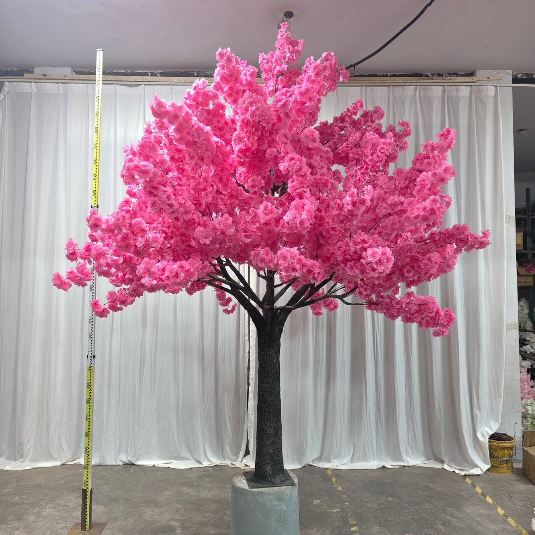 A beautiful artificial dark pink cherry blossom tree with a sturdy trunk, set on a cement base for stability, against a white curtain backdrop.