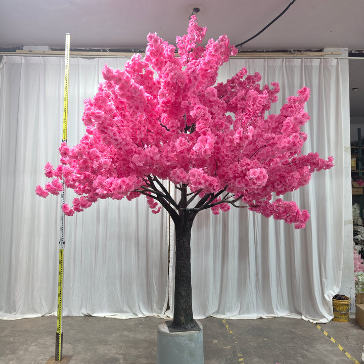 A beautiful artificial dark pink cherry blossom tree with a sturdy trunk, set on a cement base for stability, against a white curtain backdrop.