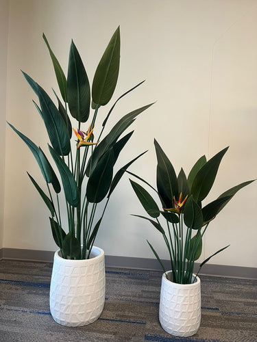 Two tall white planters with a geometric textured design, each holding artificial bird of paradise plants with vibrant green leaves and orange flowers, placed indoors on a carpeted floor. Are these planters suitable for tall plants?