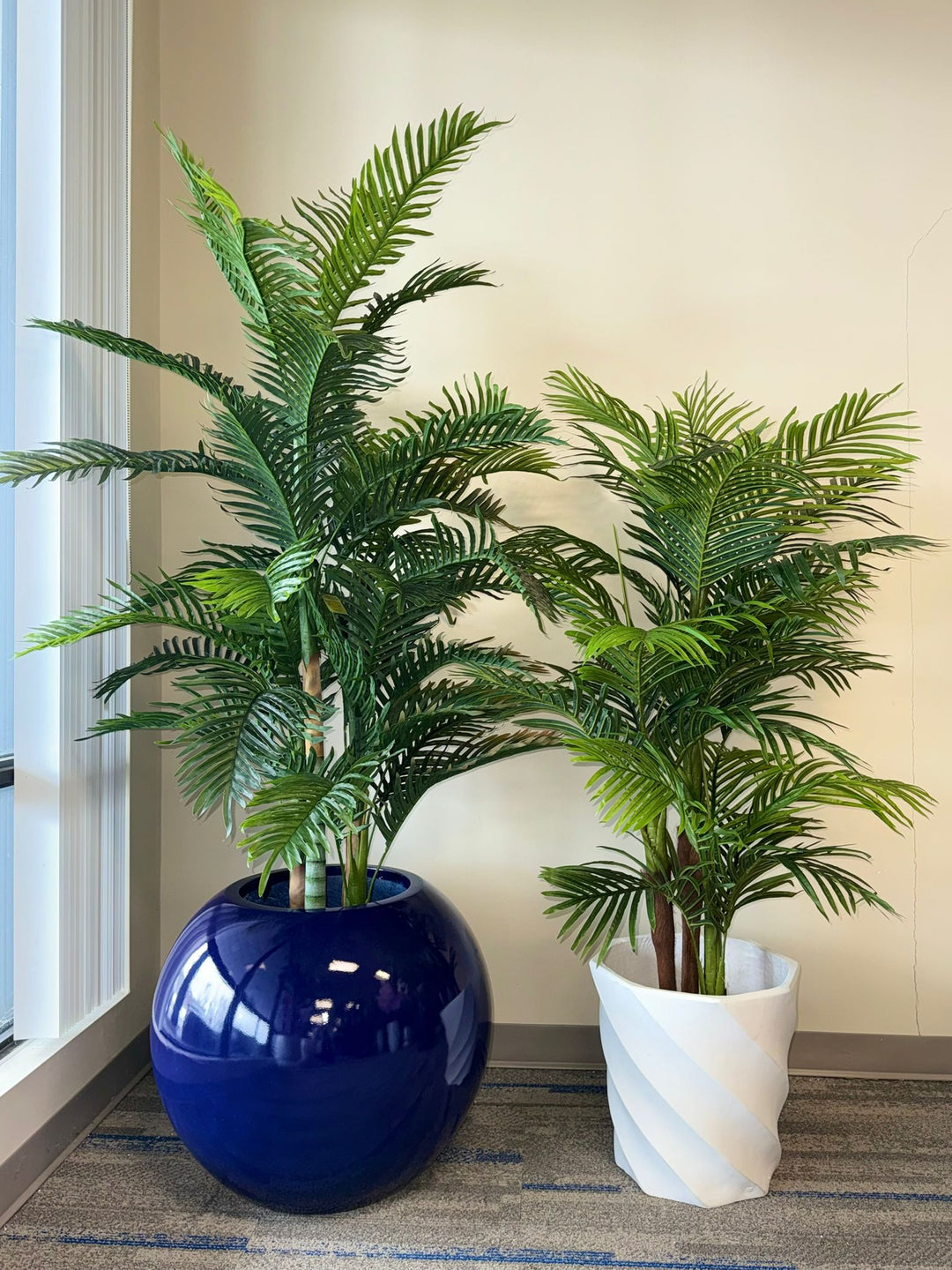 Two potted artificial tropical plants, one in a blue pot and one in a white pot, against a neutral wall.